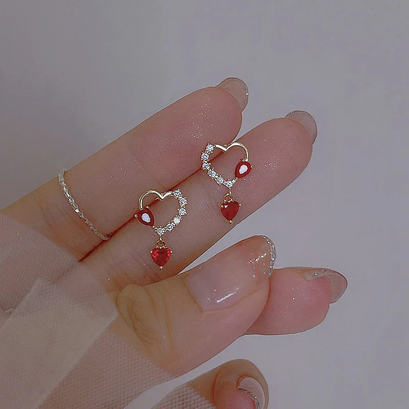 Heart-shaped earrings with red stones held between fingers against a neutral background